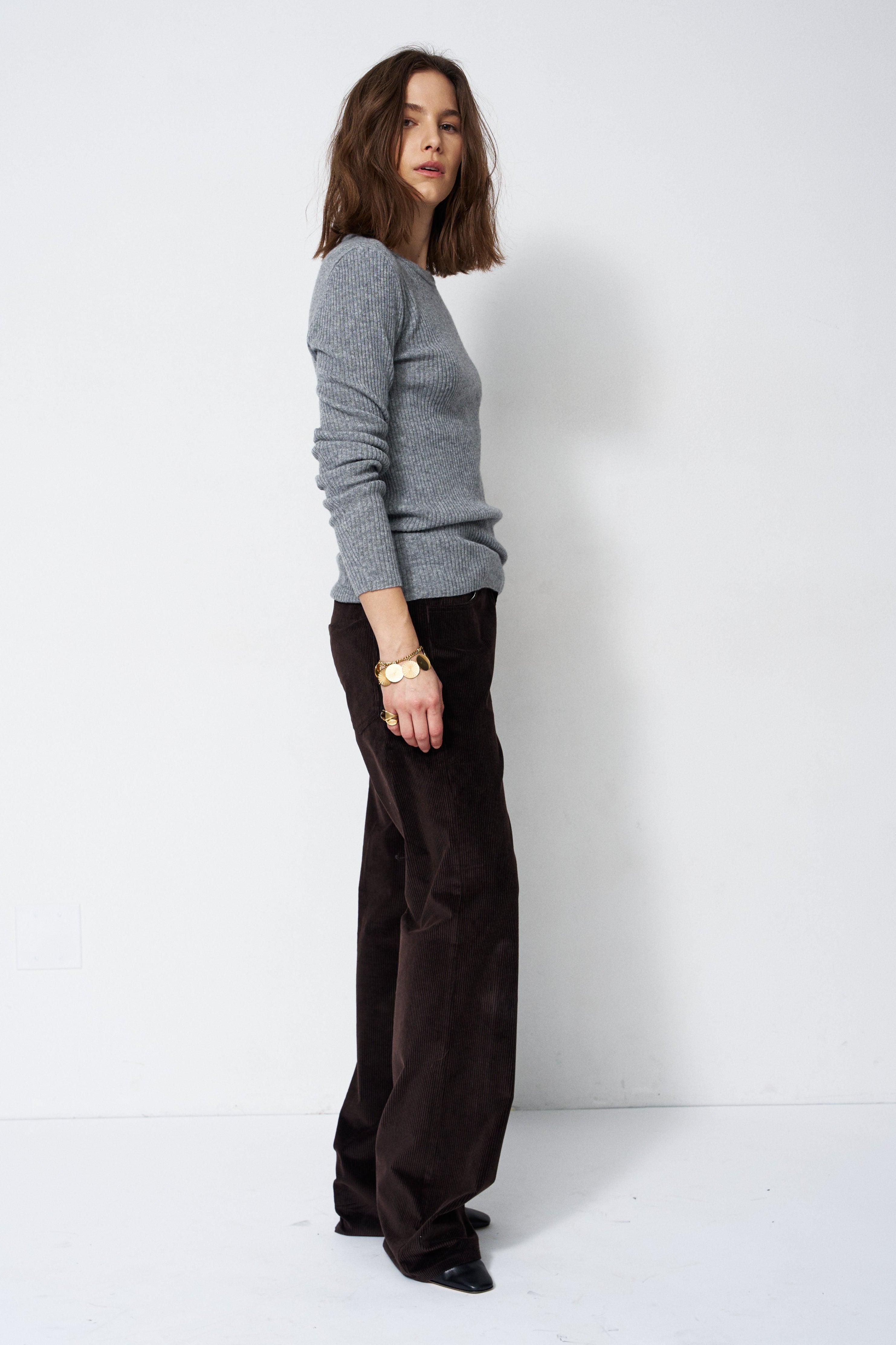 A woman stands in profile wearing a light gray long-sleeve knit sweater and dark brown wide-leg trousers against a plain white studio background.