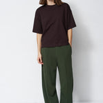model in green pants and brown shirt standing in front of a white wall
