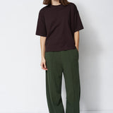model in green pants and brown shirt standing in front of a white wall