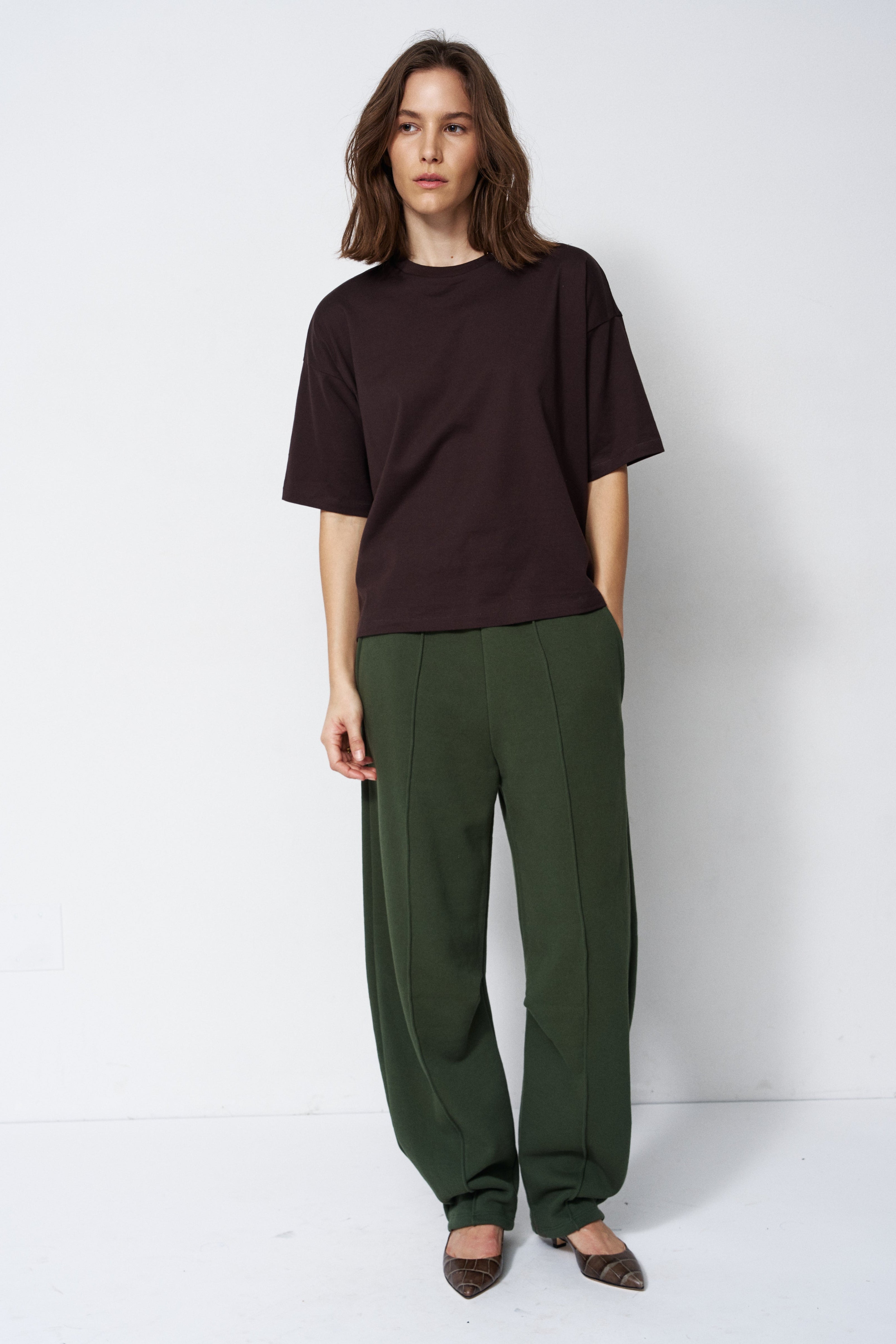 model in green pants and brown shirt standing in front of a white wall