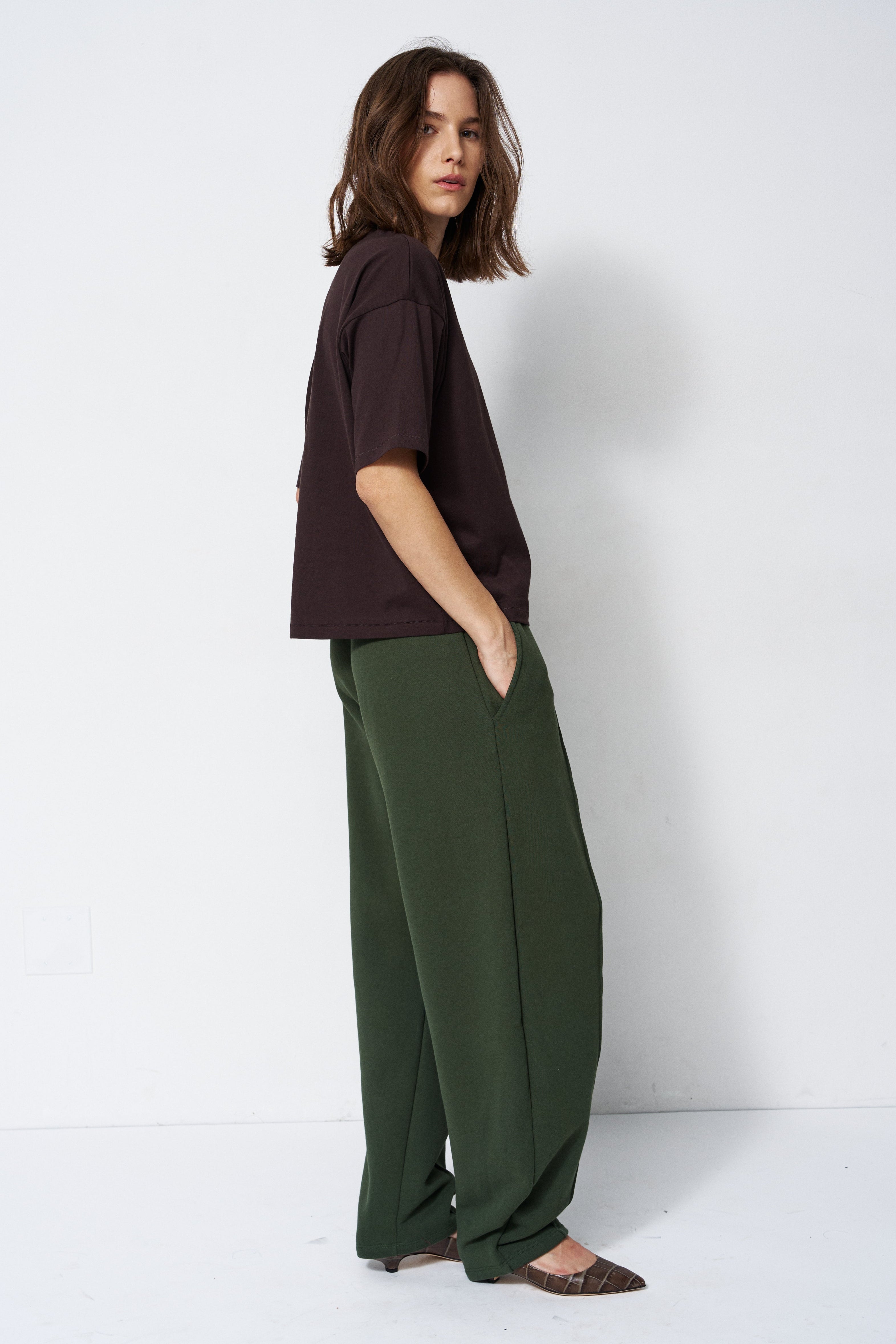 model in green pants and brown shirt standing in front of a white wall