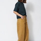 A woman stands in a studio wearing a dark green short-sleeve t-shirt and wide-leg mustard yellow trousers, paired with brown shoes.