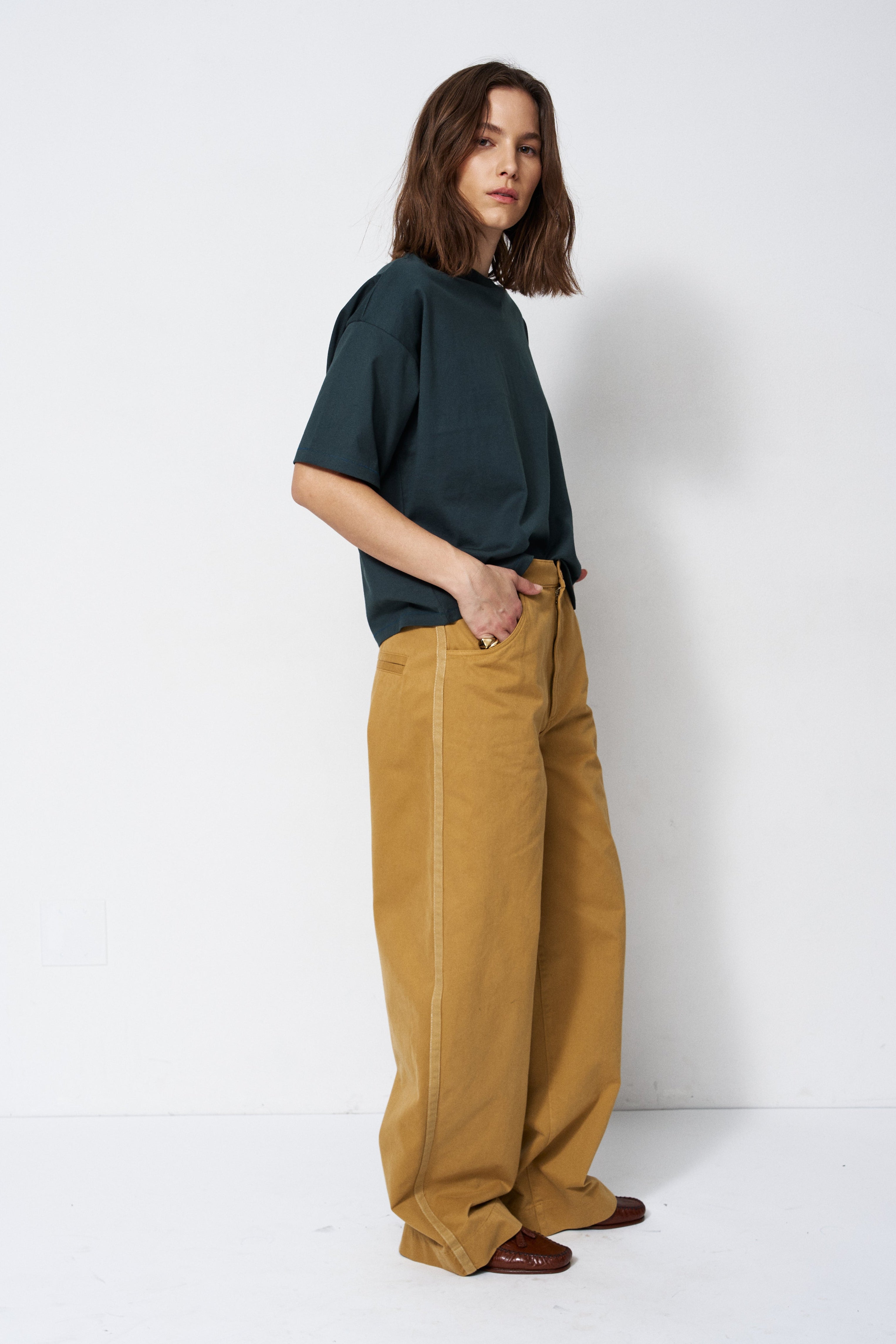A woman stands in a studio wearing a dark green short-sleeve t-shirt and wide-leg mustard yellow trousers, paired with brown shoes.