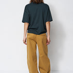 A model viewed from the back wearing an oversized dark green t-shirt and wide-leg mustard yellow pants, standing against a plain white studio background.