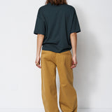 A model viewed from the back wearing an oversized dark green t-shirt and wide-leg mustard yellow pants, standing against a plain white studio background.