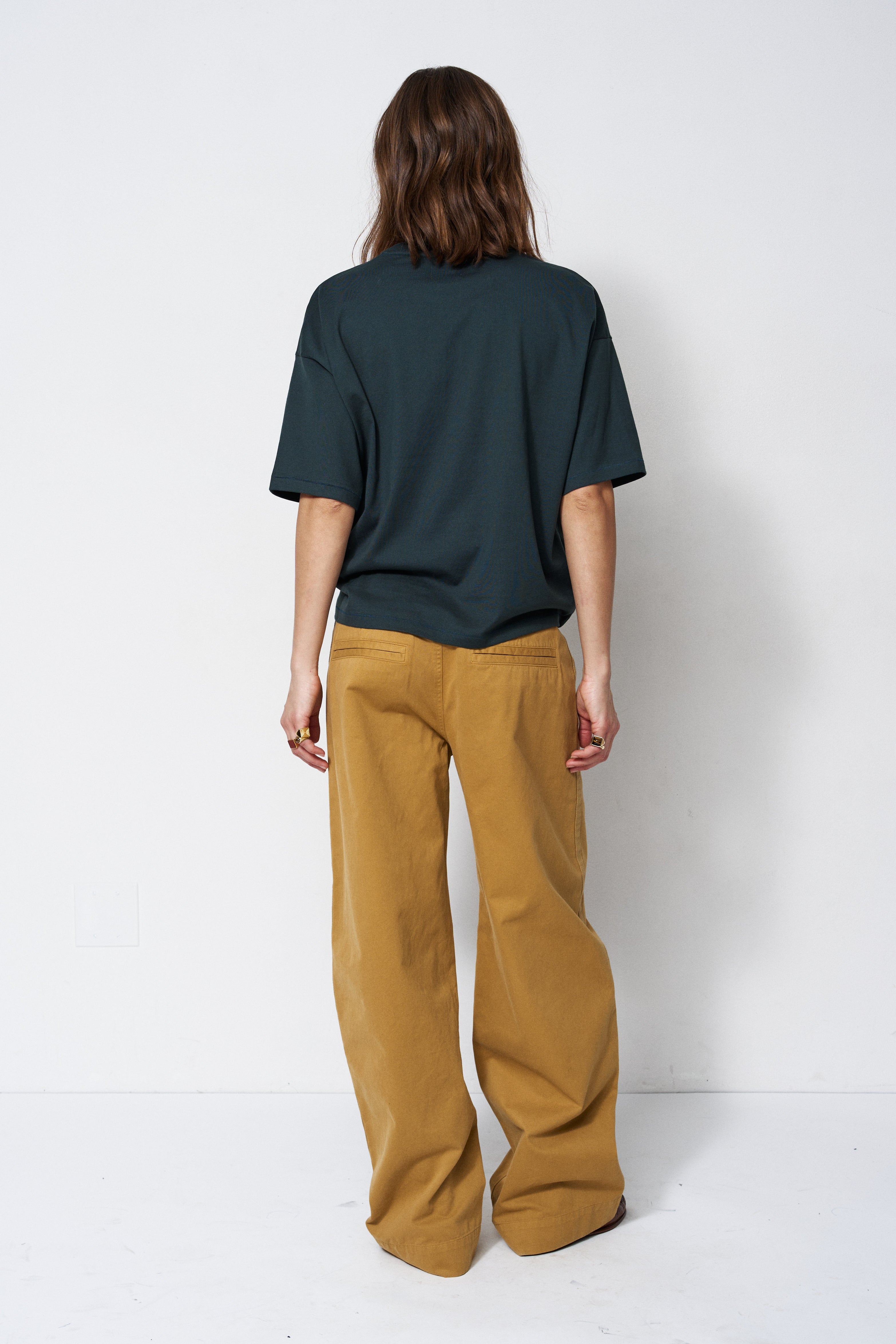 A model viewed from the back wearing an oversized dark green t-shirt and wide-leg mustard yellow pants, standing against a plain white studio background.