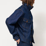 A model wearing a dark blue denim jacket and matching dark blue denim pants, paired with brown suede ankle boots, standing in profile against a plain white studio background.