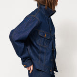 A model wearing a dark blue denim jacket and matching dark blue denim pants, paired with brown suede ankle boots, standing in profile against a plain white studio background.
