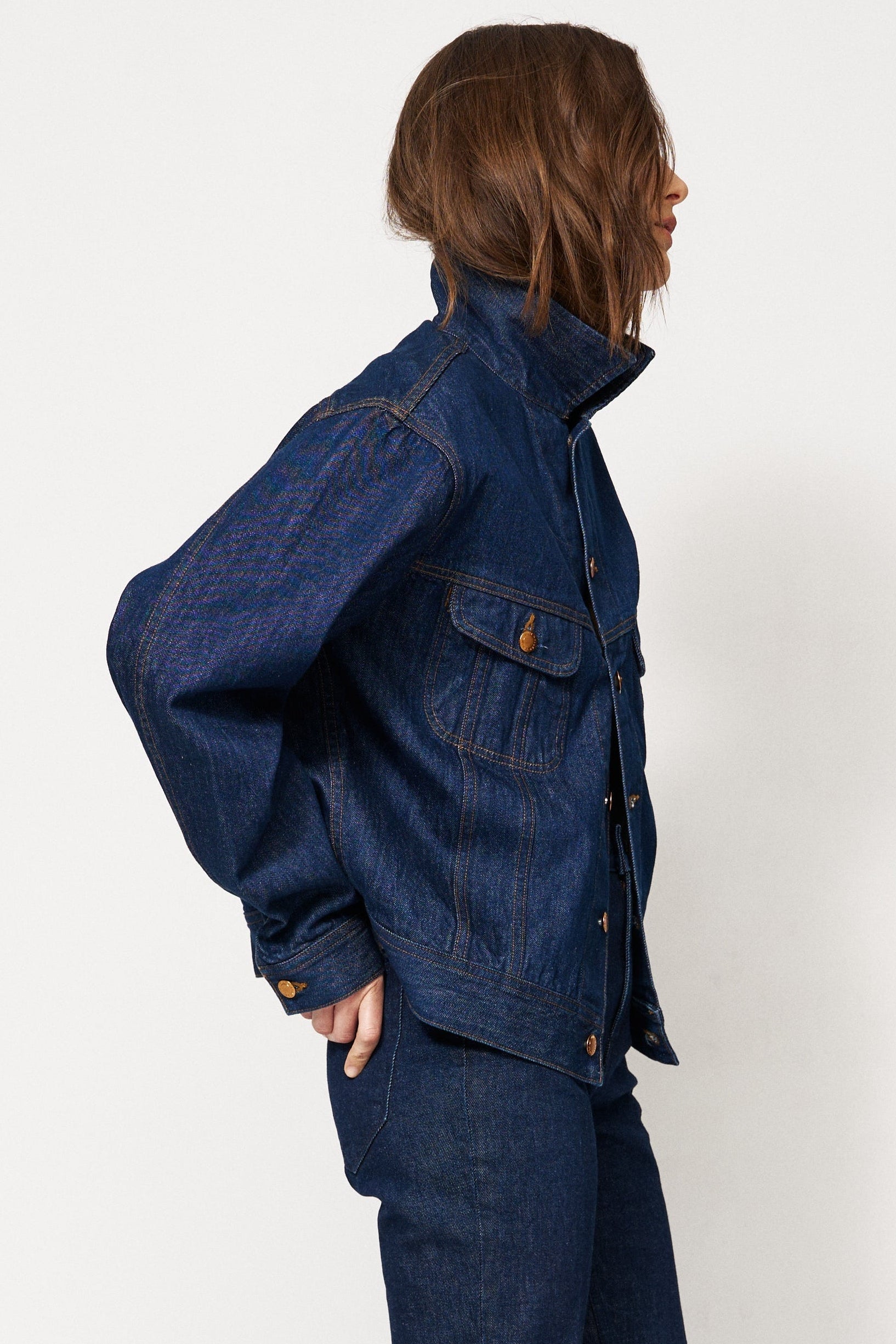A model wearing a dark blue denim jacket and matching dark blue denim pants, paired with brown suede ankle boots, standing in profile against a plain white studio background.