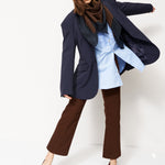 A woman modeling a stylish outfit consisting of a navy blue oversized blazer, a light blue shirt, a dark brown scarf, brown wide-leg pants, and burgundy pointed-toe heels, set against a plain white studio background.