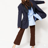 A woman modeling a stylish outfit consisting of a navy blue oversized blazer, a light blue shirt, a dark brown scarf, brown wide-leg pants, and burgundy pointed-toe heels, set against a plain white studio background.
