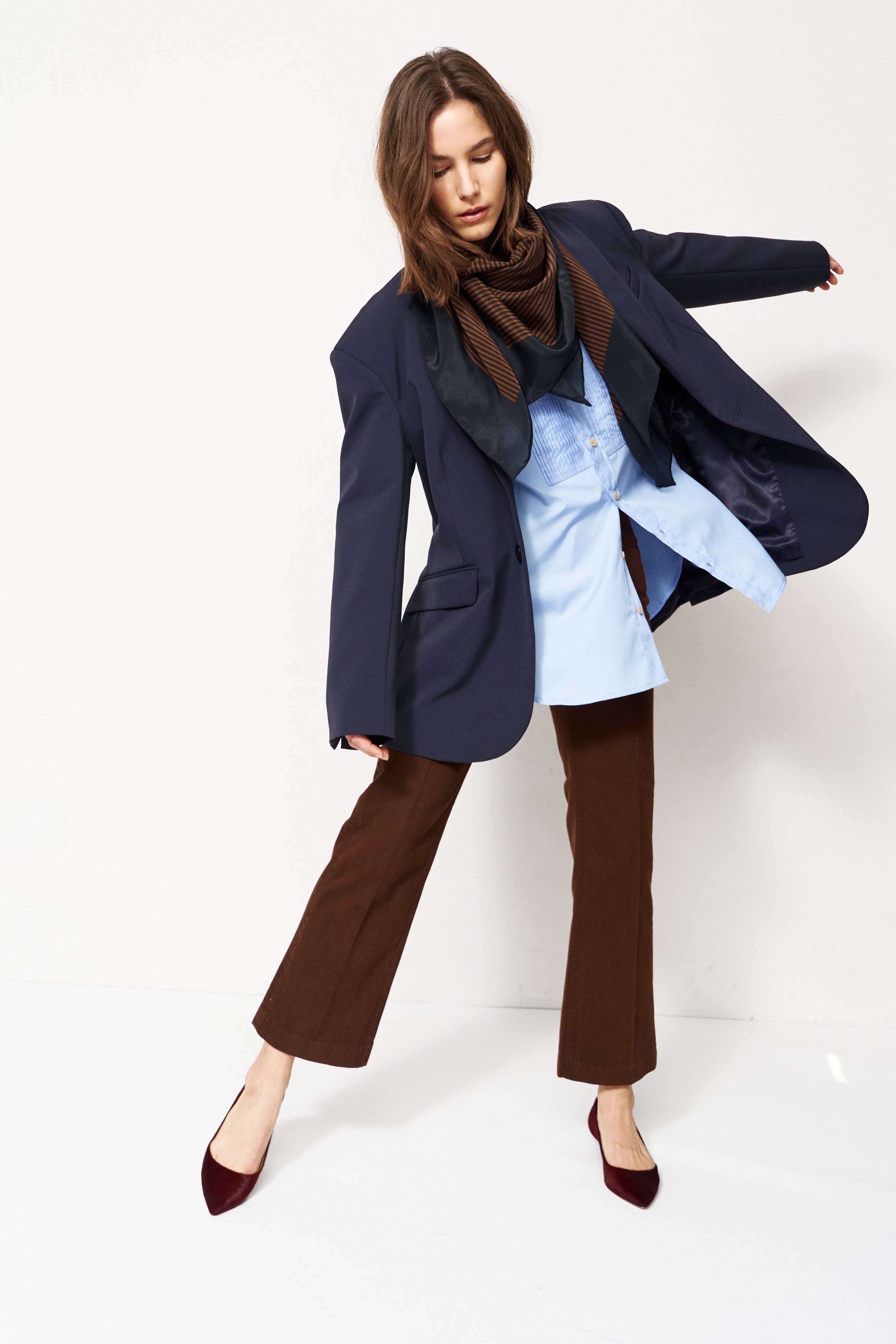 A woman modeling a stylish outfit consisting of a navy blue oversized blazer, a light blue shirt, a dark brown scarf, brown wide-leg pants, and burgundy pointed-toe heels, set against a plain white studio background.