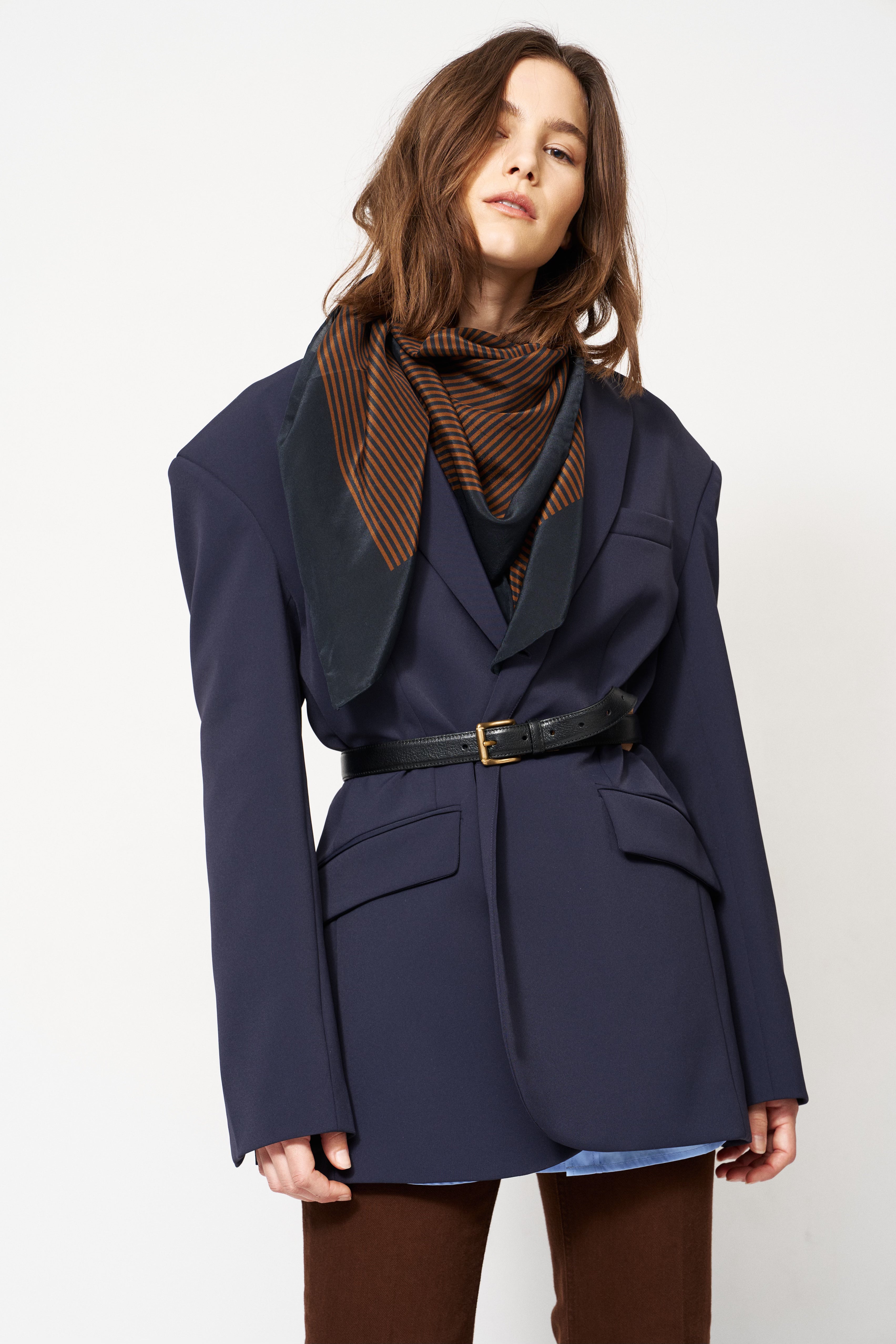 A model wearing a navy blue oversized blazer with a black belt, a brown and black striped scarf, and brown trousers, photographed against a plain white studio background.