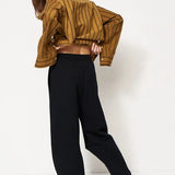 woman in gold blouse and black trackpant in front of a white wall