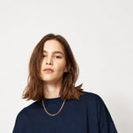 A woman with shoulder-length brown hair wearing a dark navy oversized crewneck sweatshirt and a delicate gold chain necklace, photographed against a solid light gray background.