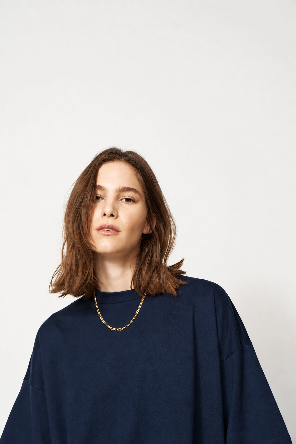 A woman with shoulder-length brown hair wearing a dark navy oversized crewneck sweatshirt and a delicate gold chain necklace, photographed against a solid light gray background.