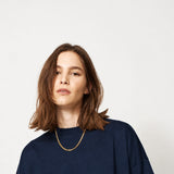 A woman with shoulder-length brown hair wearing a dark navy oversized crewneck sweatshirt and a delicate gold chain necklace, photographed against a solid light gray background.