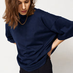A woman wearing a dark navy oversized turtleneck top and black pants, photographed against a plain light gray studio background.