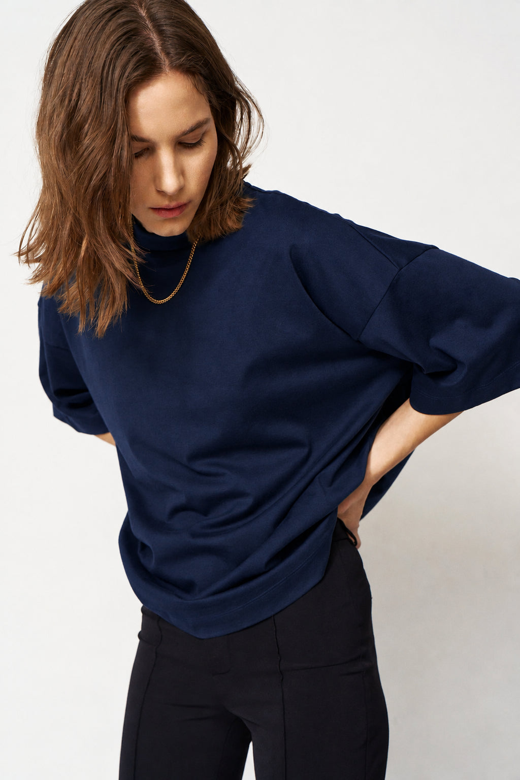 A woman wearing a dark navy oversized turtleneck top and black pants, photographed against a plain light gray studio background.