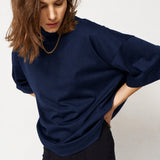 A woman wearing a dark navy oversized turtleneck top and black pants, photographed against a plain light gray studio background.