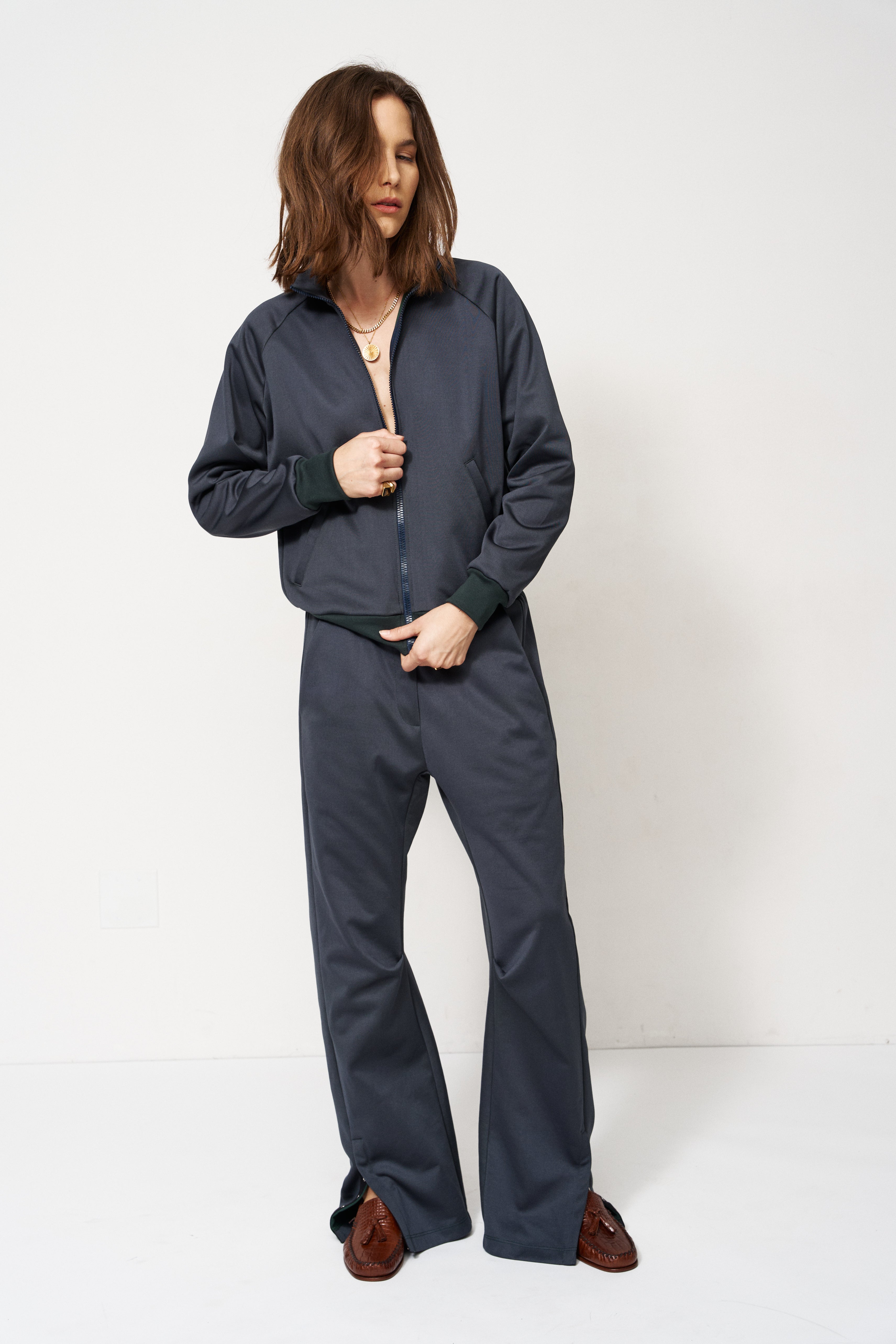 A woman modeling a dark navy blue zip-up jacket and matching wide-leg pants set, paired with brown shoes, against a plain white studio background.