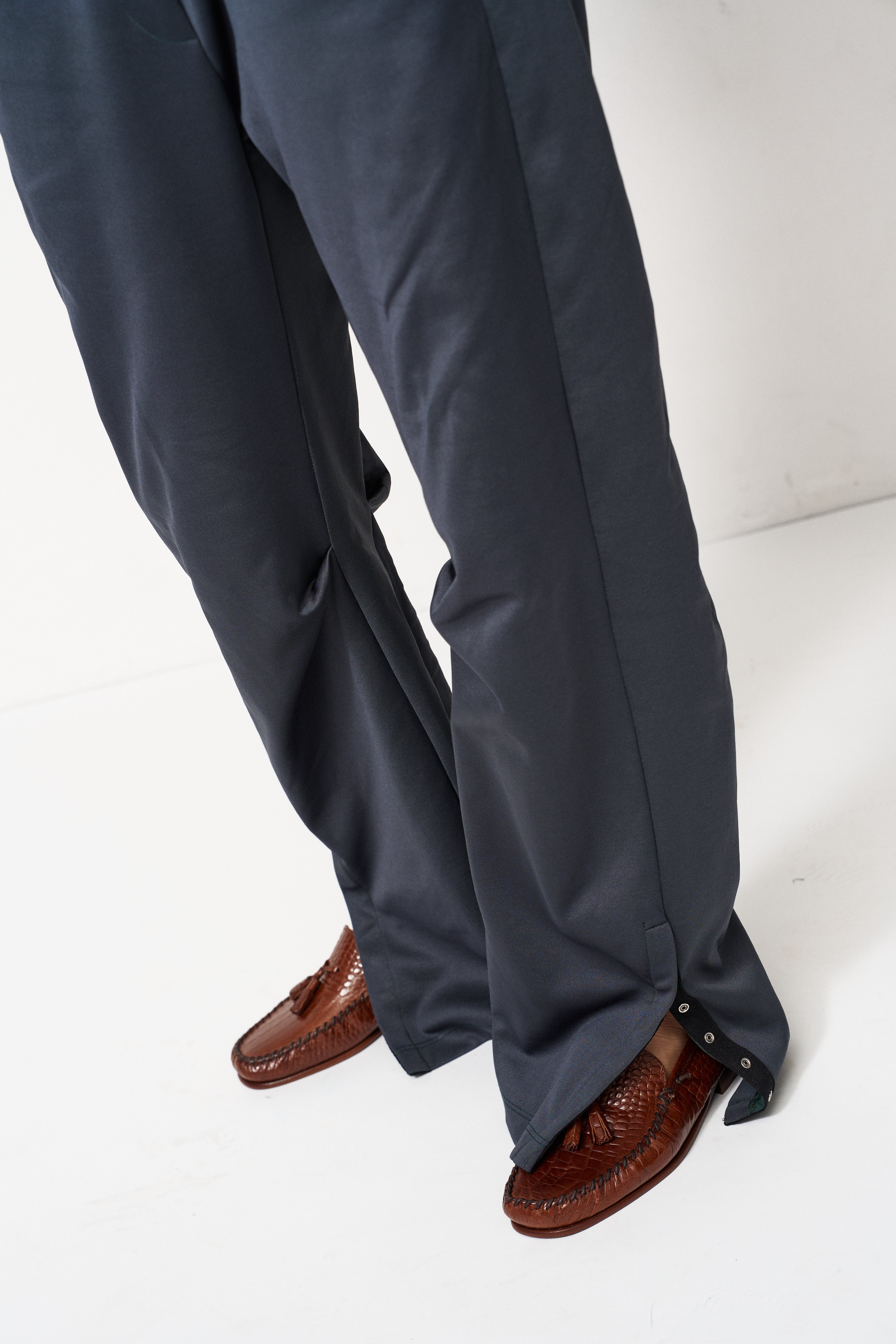 Close-up of a person wearing dark charcoal gray wide-leg trousers with side buttons and brown leather loafers, shown from the knees down against a white studio background.