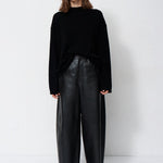 A woman stands against a plain white studio background wearing a black long-sleeve mock-neck top, black wide-leg leather pants, and black pointed ankle boots.