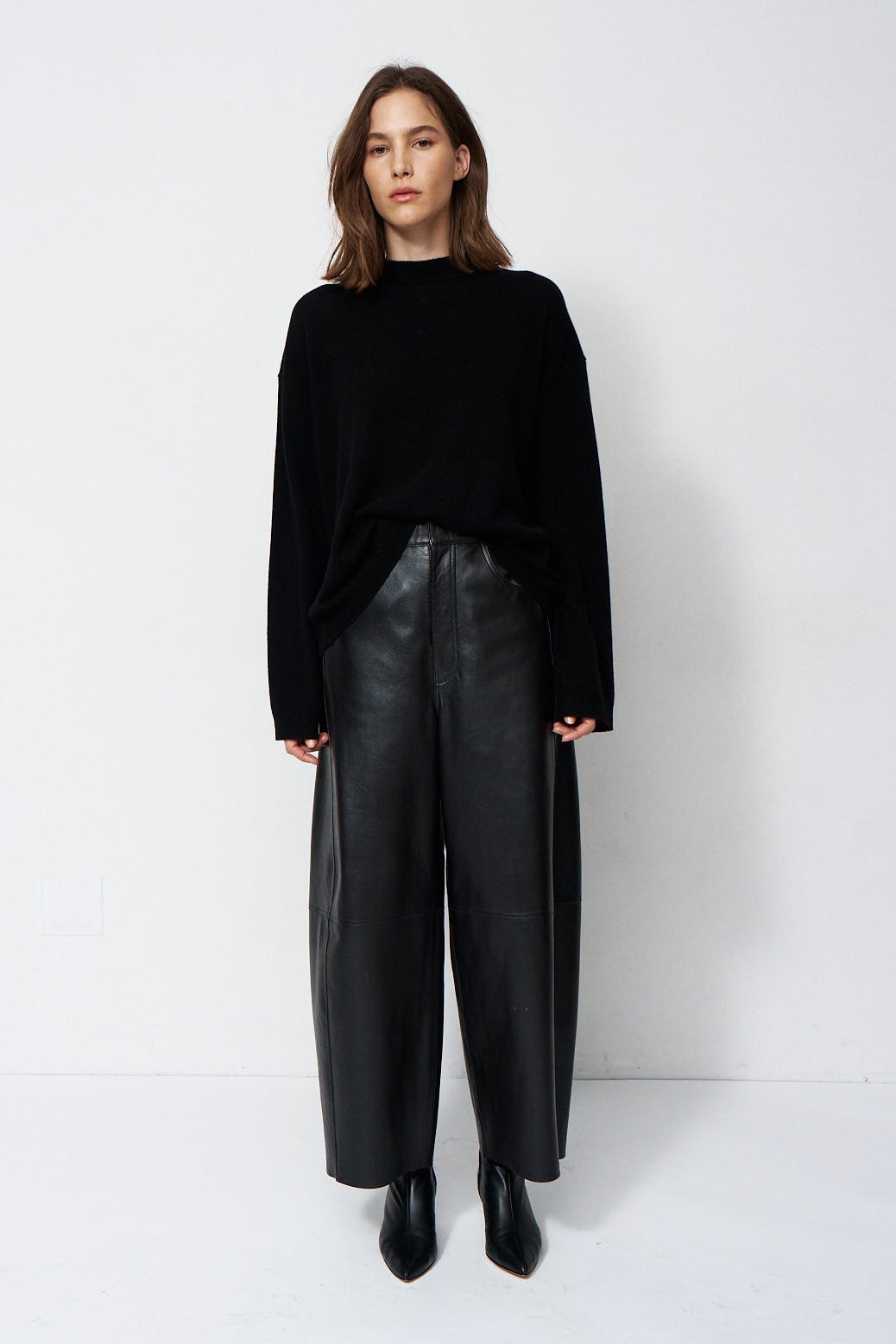 A woman stands against a plain white studio background wearing a black long-sleeve mock-neck top, black wide-leg leather pants, and black pointed ankle boots.