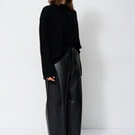 A woman modeling a black long-sleeve knit top and black wide-leg leather pants against a plain white studio background.