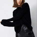 A woman modeling a black long-sleeve knit top with a relaxed, slightly oversized fit, paired with black leather pants, against a plain light gray studio background.