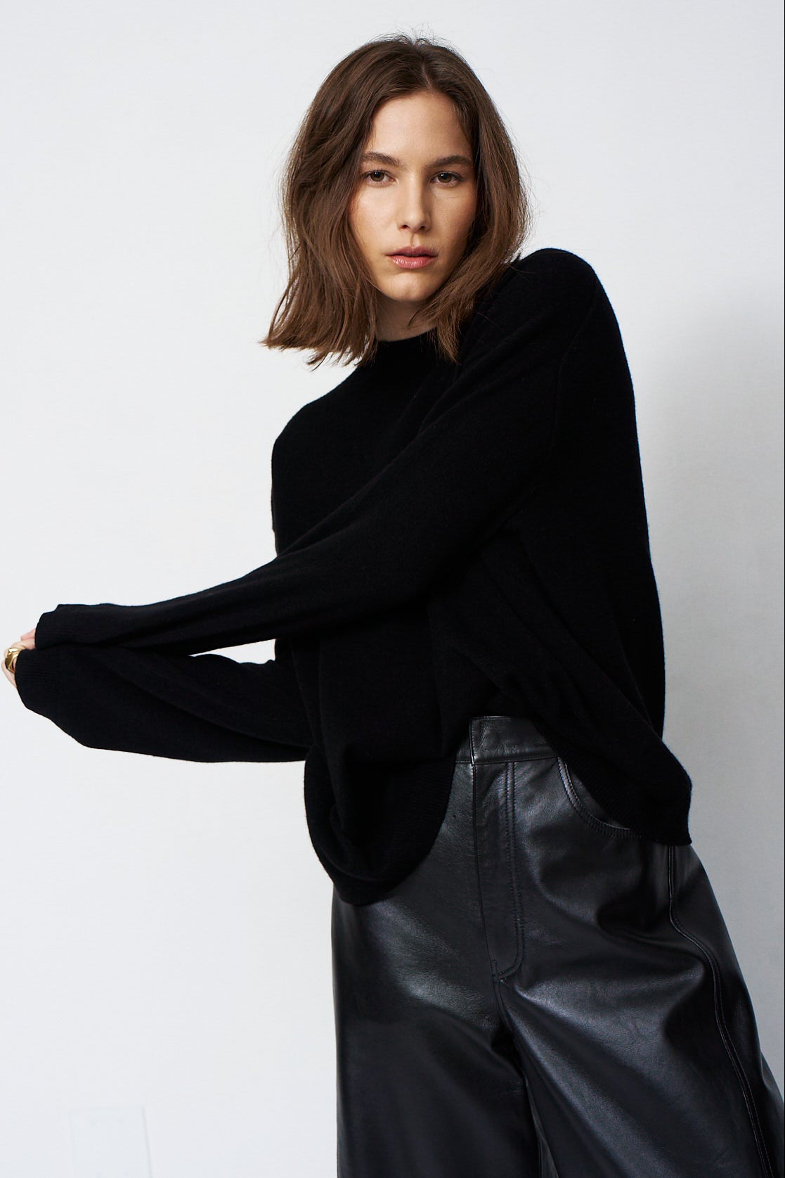 A woman modeling a black long-sleeve knit top with a relaxed, slightly oversized fit, paired with black leather pants, against a plain light gray studio background.