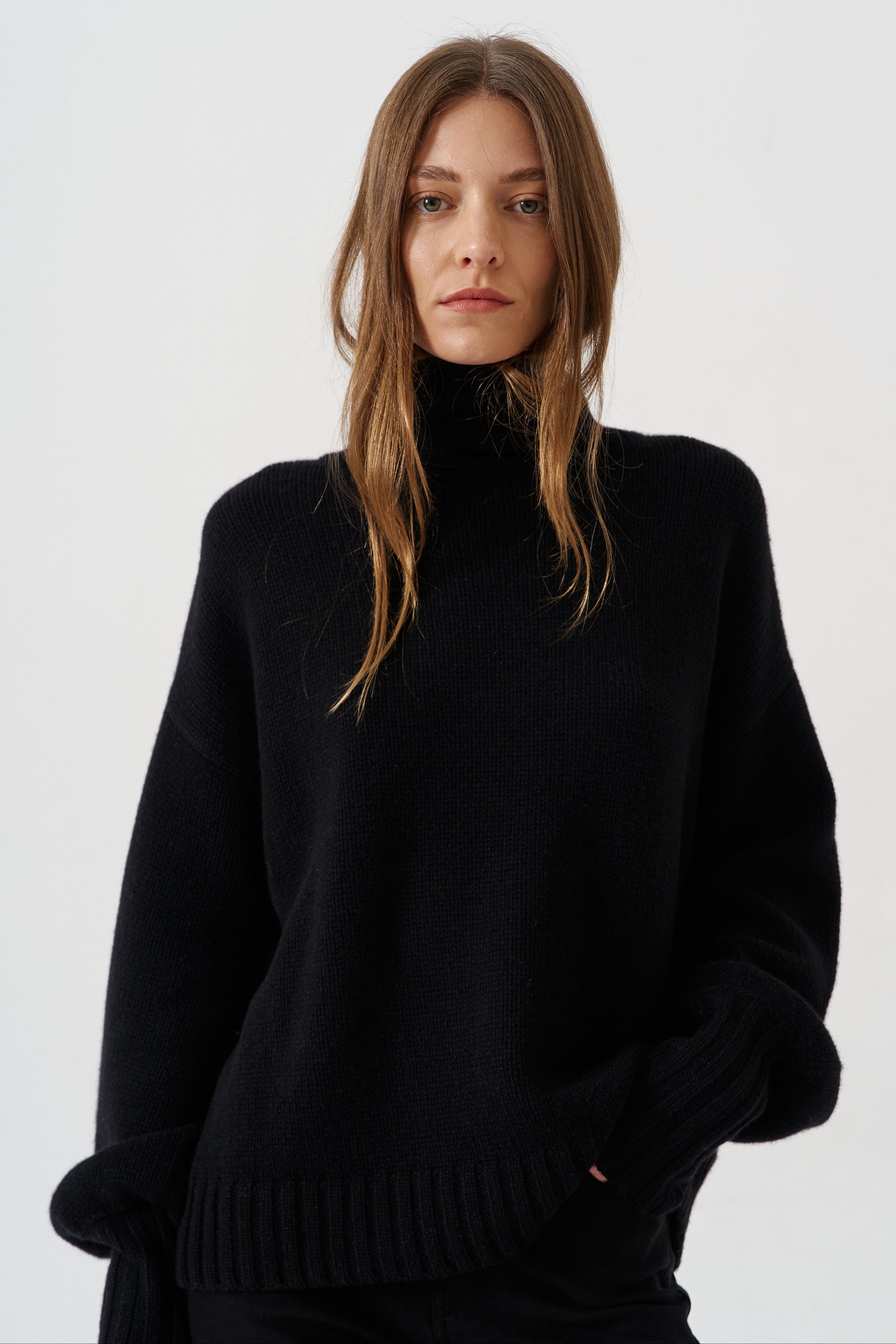 A woman wearing an oversized black knit turtleneck sweater, photographed against a clean light gray studio background.