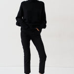 A model stands against a plain white studio background wearing a black turtleneck sweater, black cropped trousers, and bright red pointed-toe heels.