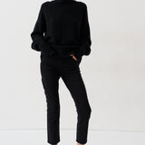 A model stands against a plain white studio background wearing a black turtleneck sweater, black cropped trousers, and bright red pointed-toe heels.