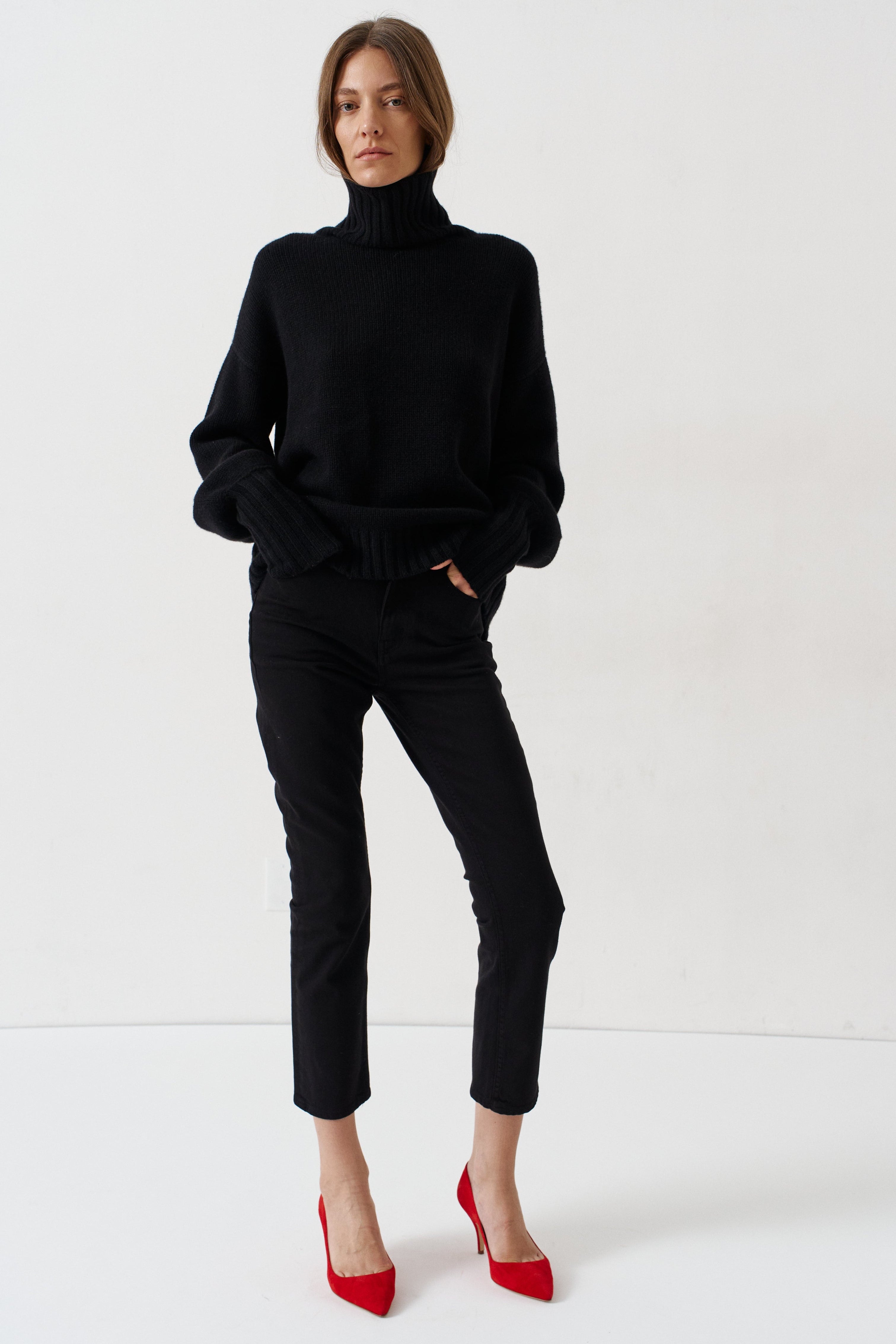 A model stands against a plain white studio background wearing a black turtleneck sweater, black cropped trousers, and bright red pointed-toe heels.