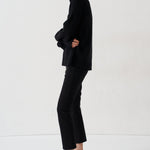 A woman stands in profile wearing a black turtleneck sweater, black cropped trousers, and bright red pointed-toe high heels against a clean white studio background.