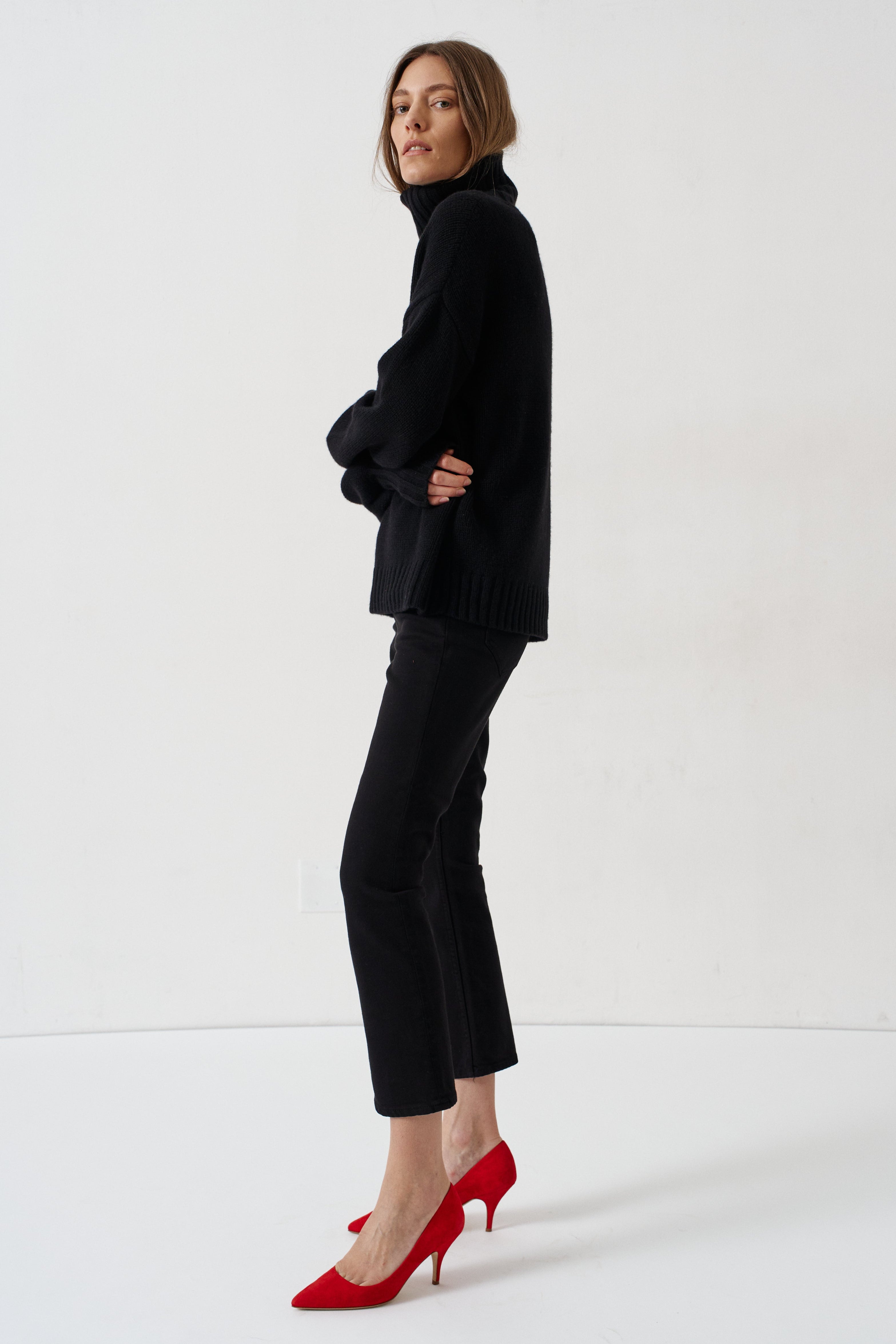 A woman stands in profile wearing a black turtleneck sweater, black cropped trousers, and bright red pointed-toe high heels against a clean white studio background.