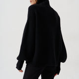 A woman wearing a black oversized knit sweater with a high ribbed turtleneck and long balloon sleeves, shown from the back against a plain light gray studio background.