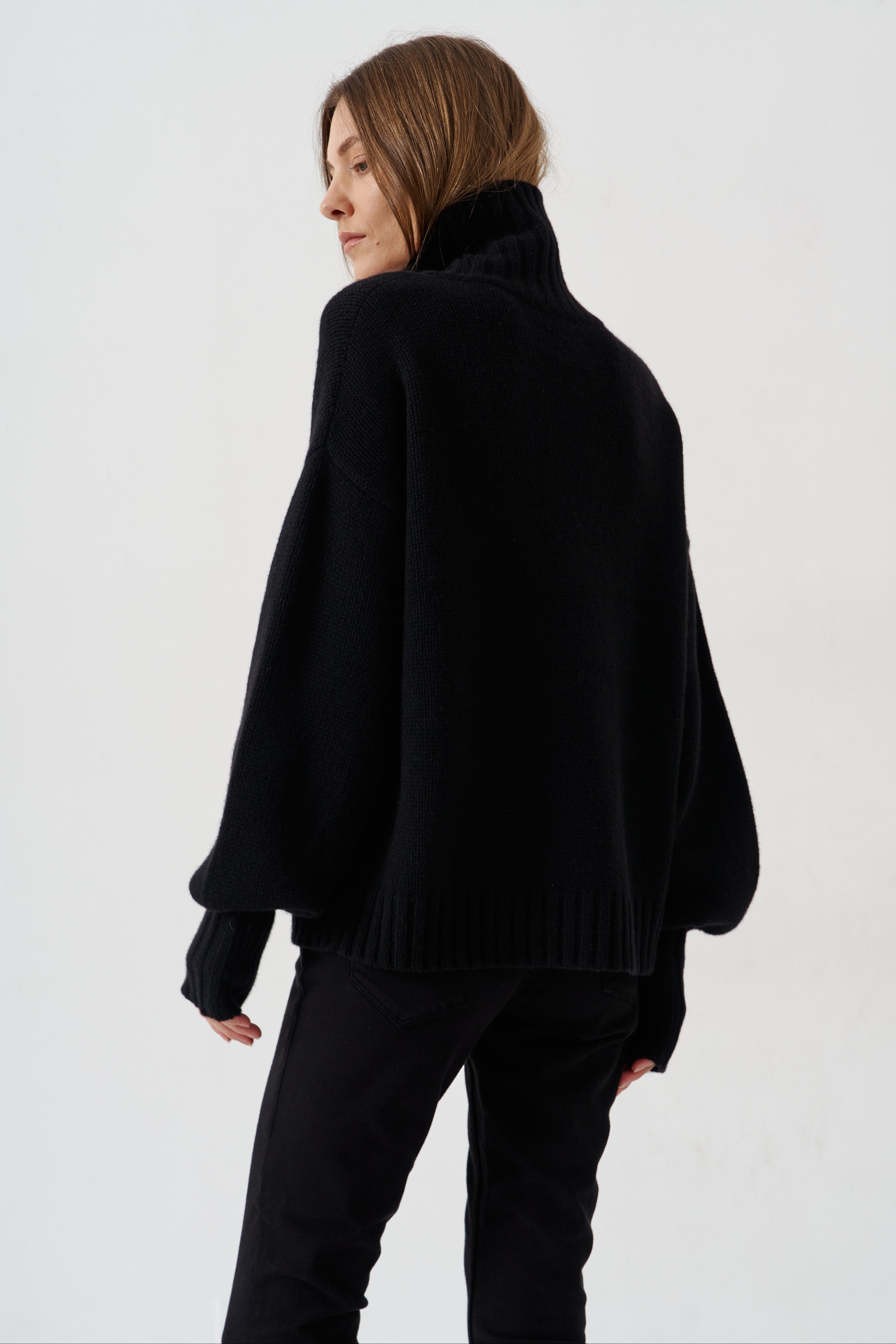A woman wearing a black oversized knit sweater with a high ribbed turtleneck and long balloon sleeves, shown from the back against a plain light gray studio background.