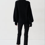 A model viewed from behind wearing a black oversized turtleneck sweater, black cropped trousers, and bright red pointed-toe heels against a plain white studio background.