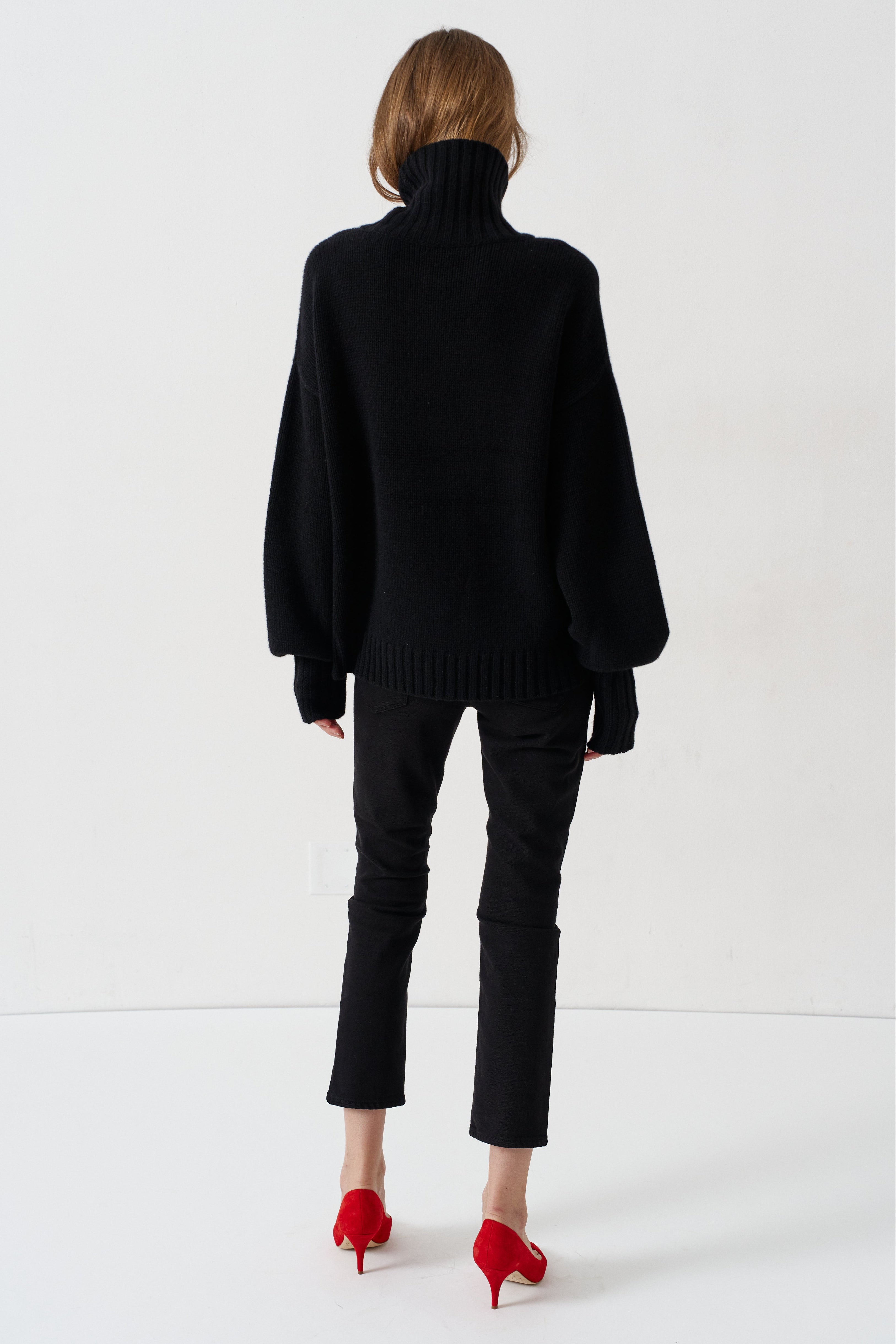 A model viewed from behind wearing a black oversized turtleneck sweater, black cropped trousers, and bright red pointed-toe heels against a plain white studio background.