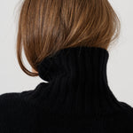 Rear view of a person with shoulder-length brown hair wearing a black ribbed turtleneck sweater against a plain light gray background.