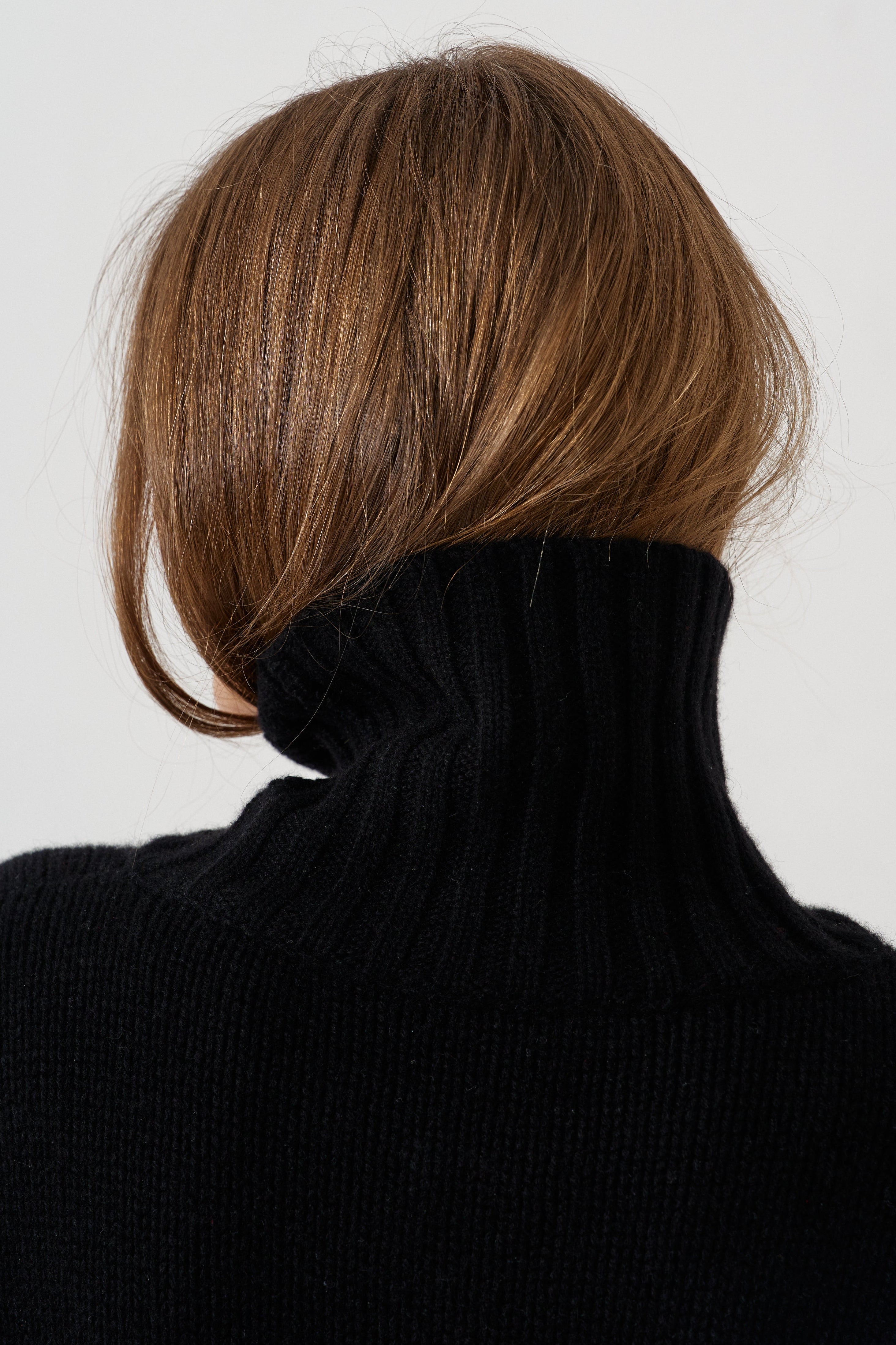 Rear view of a person with shoulder-length brown hair wearing a black ribbed turtleneck sweater against a plain light gray background.