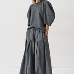 A woman modeling a dark gray two-piece outfit consisting of a loose short-sleeve top with voluminous puff sleeves and matching wide-leg pleated trousers, shot against a plain white studio background.