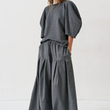 A woman modeling a dark gray two-piece outfit consisting of a loose short-sleeve top with voluminous puff sleeves and matching wide-leg pleated trousers, shot against a plain white studio background.