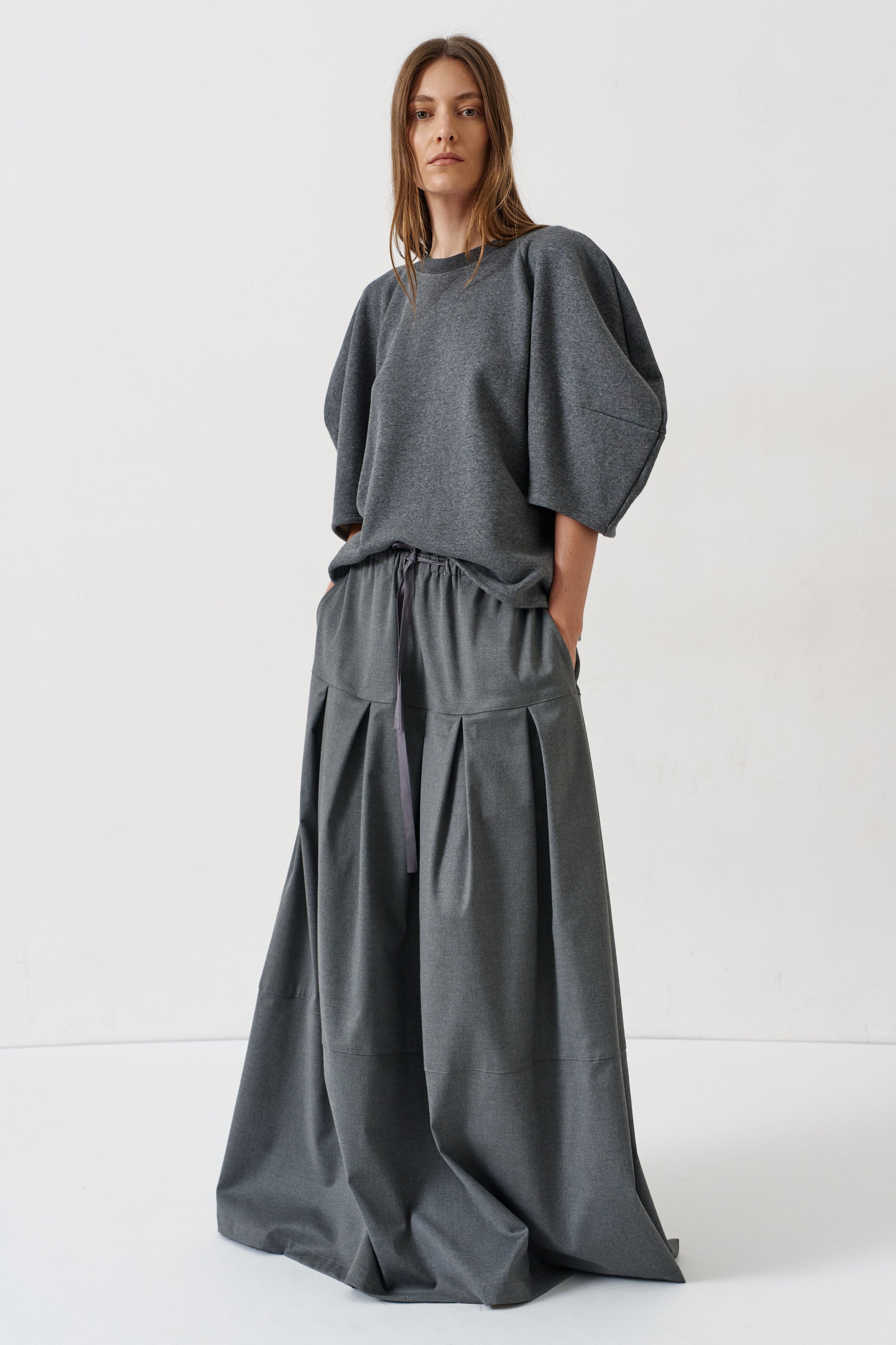 A woman modeling a dark gray two-piece outfit consisting of a loose short-sleeve top with voluminous puff sleeves and matching wide-leg pleated trousers, shot against a plain white studio background.