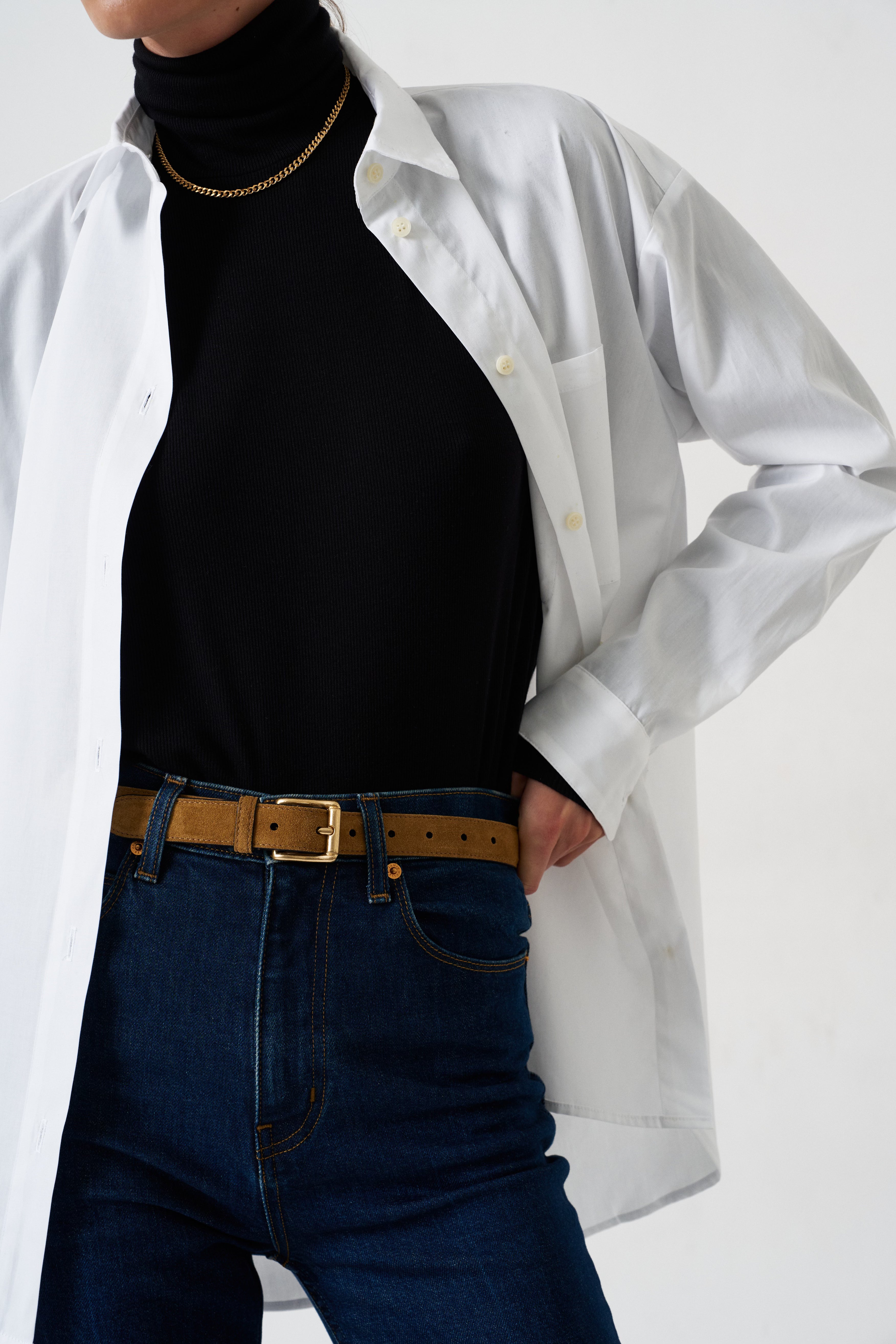 A person wearing an oversized white button-up shirt over a black turtleneck, paired with dark blue jeans and a tan belt, against a plain light background.