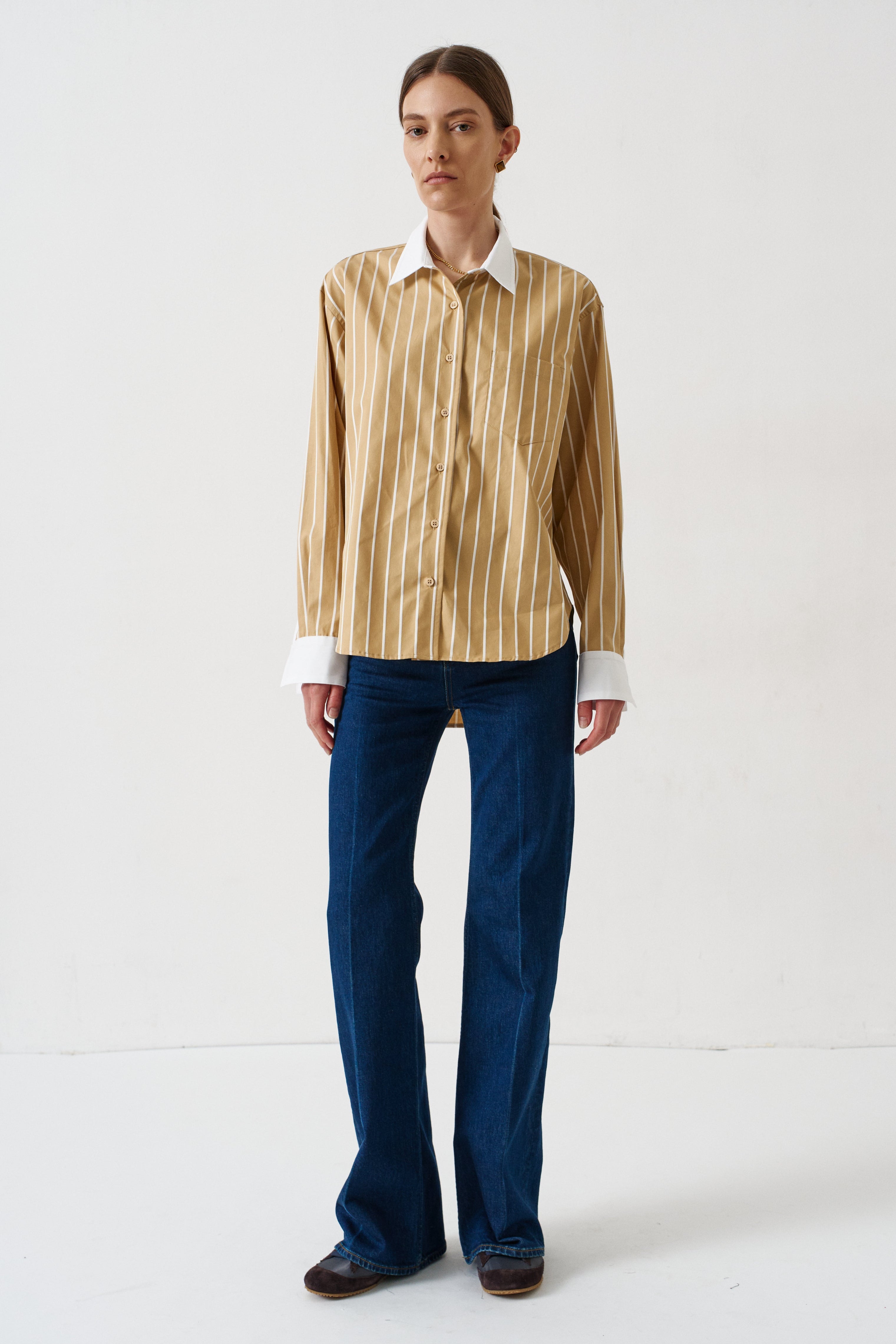 woman in gold striped shirt and jeans againsta a white wall 