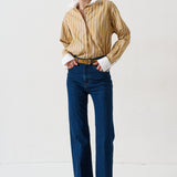woman in gold striped shirt and jeans againsta a white wall 