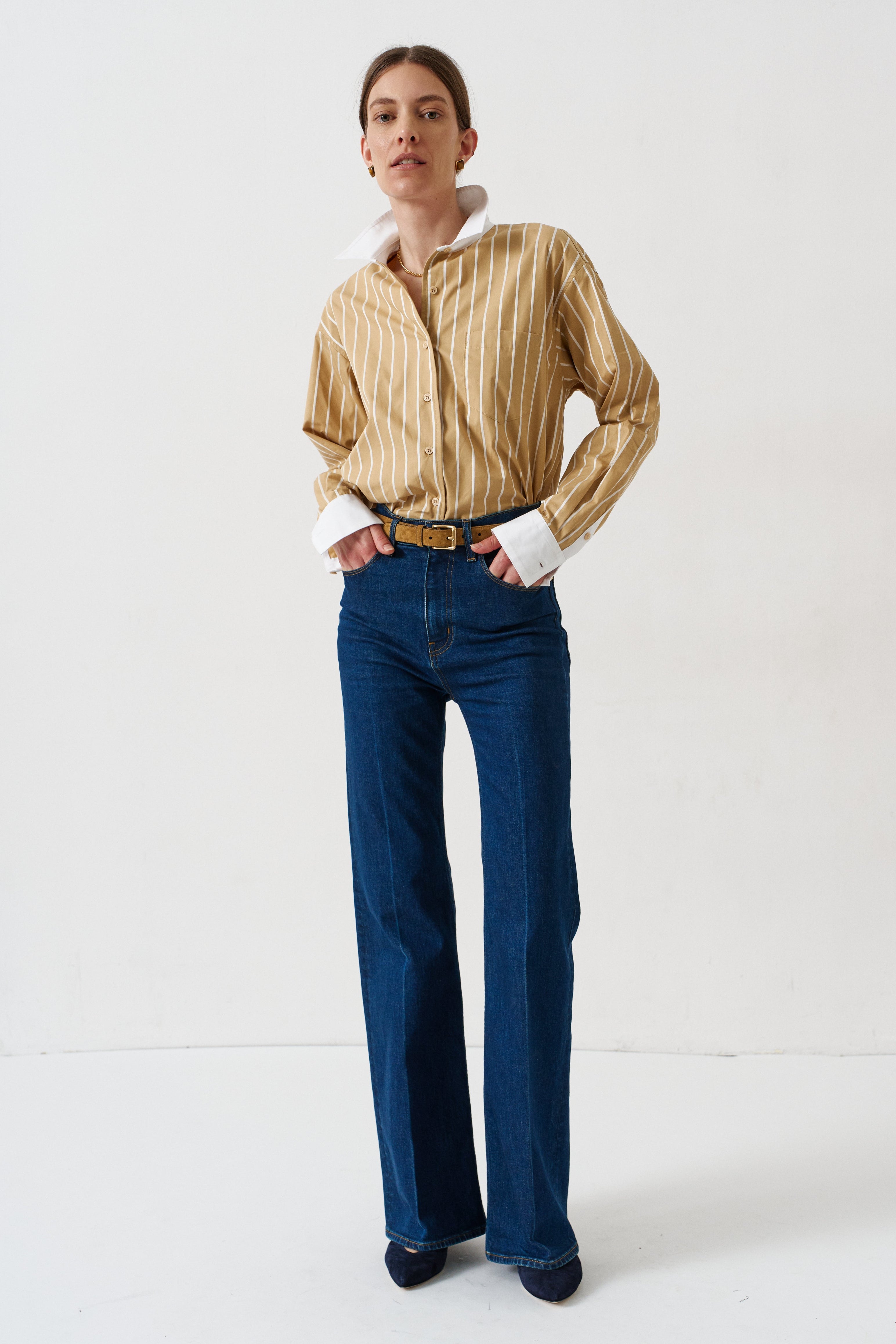 woman in gold striped shirt and jeans againsta a white wall 